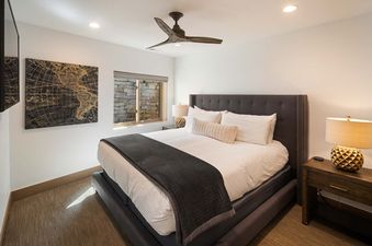 A luxurious bedroom at Silver Lode Escape features a large bed with a dark gray headboard, white linens, and a dark gray quilted throw, illuminated by recessed lighting and natural light from a window revealing a stone wall.
