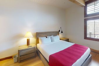 The bedroom at Manor Vail 360 features a king bed with a red comforter, two nightstands, and a large window with a view of the mountains.