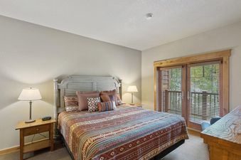 The bedroom features a queen-sized bed with a distressed white wooden headboard and a colorful patterned comforter, flanked by wooden nightstands with lamps, and a glass door leading to a balcony.