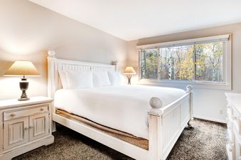 A serene bedroom features a white wooden bed with crisp white linens, flanked by nightstands with lamps, and a large window overlooking autumn trees with a dusting of snow.