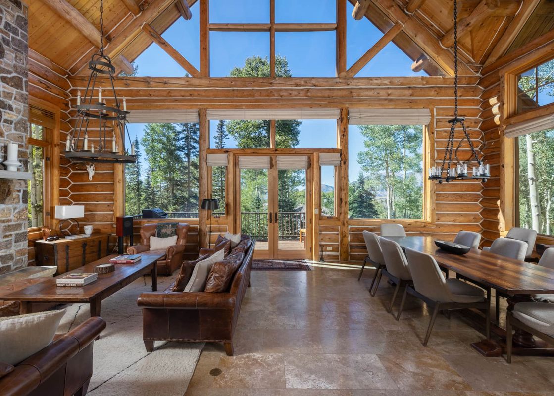 The Bear Lodge's great room features a living area with leather furniture, a stone fireplace, and a dining area with a long wooden table, all set against a backdrop of large windows offering mountain and forest views.