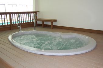 An indoor hot tub with bubbling water sits on a light wooden deck, featuring a small wooden bench nearby and a railing in the background.