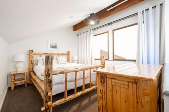 A bright bedroom at Snow Flower 129 Park City features a king-sized log bed with white bedding, two large windows with white curtains, and a ceiling fan.