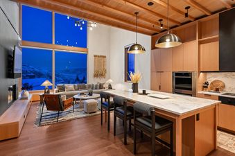 A wide view of the combined living and kitchen area at Havens Summit, featuring a cozy sofa, coffee table, leather lounge chair, and a sleek fireplace, adjacent to a kitchen island with bar stools, wood cabinetry, and large windows revealing a snowy mount