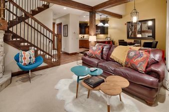 A cozy living room in Silver Star 702 with a leather sofa, blue accent chair, and coffee tables, with a staircase leading upstairs and a full kitchen visible in the background.