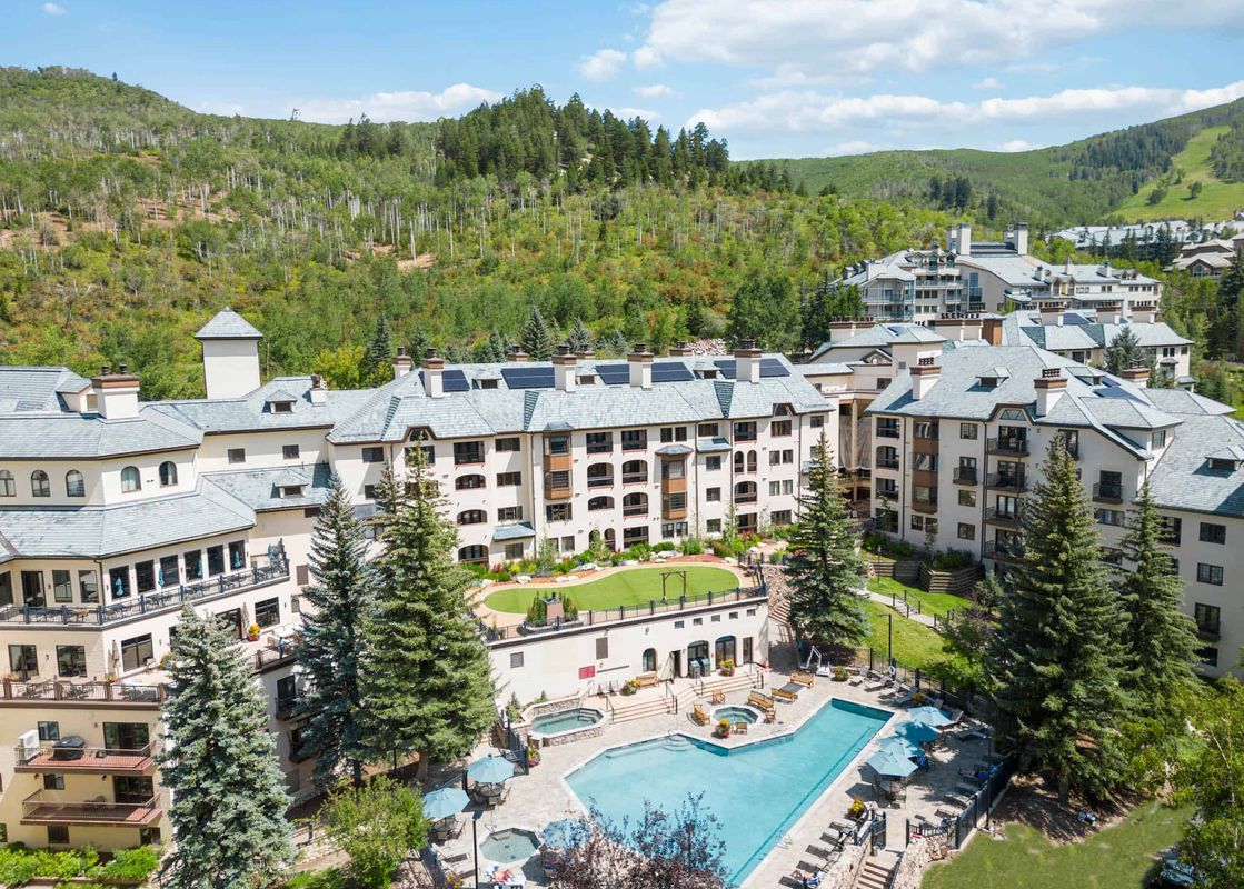 An aerial view of The Charter C1105, a multi-story building surrounded by green mountains and evergreen trees, with an outdoor pool and hot tubs.