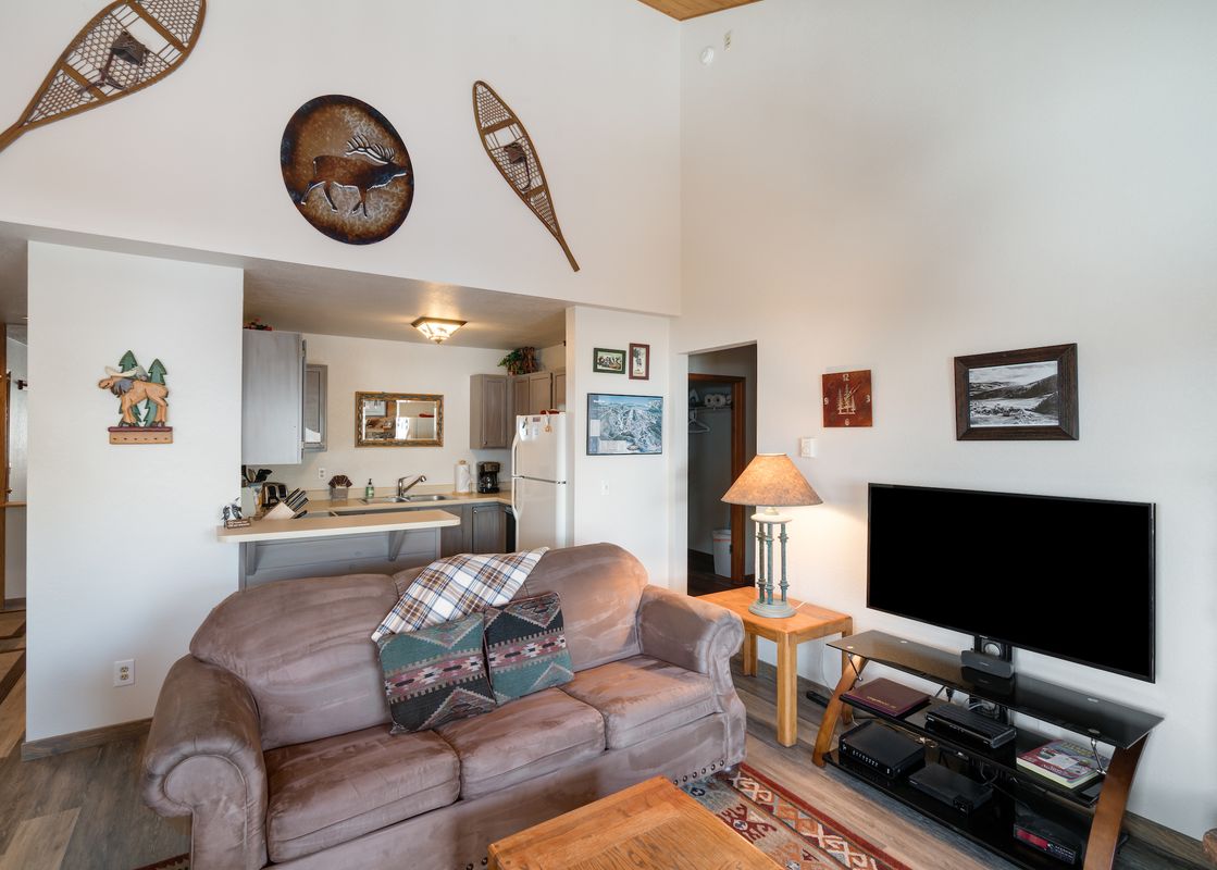 A cozy open-concept living room with a brown sofa and a kitchen area featuring modern appliances at The Pines Condominiums A304.