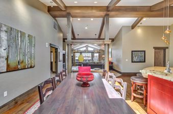 A spacious dining room with a long wooden table, multiple chairs, and a red decorative bowl in the center. In the background, there is a living area with a large window and a kitchen area with bar stools.
