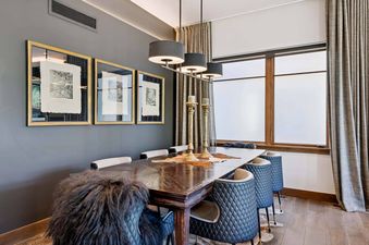 A sophisticated dining room featuring a dark wood table with a fur throw, modern black and beige quilted dining chairs, a multi-shade chandelier, framed artwork, and elegant drapes.