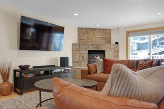 A cozy living room in Resort Plaza 5043 with a stone fireplace, large wall-mounted TV, brown leather sofas, and a view of snowy buildings outside.