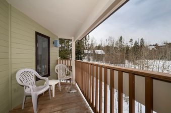 A private balcony at Silverado II Unit 334, featuring two white chairs and a small table, offers a view of a snow-covered landscape with trees and distant buildings under a cloudy sky.