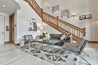 The Silver Strike Lodge 304 features a grand living room with a large gray sectional sofa, two armchairs, a coffee table, and an area rug, beneath a stunning wooden staircase.