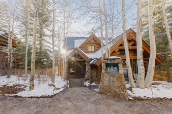 The Frostpine Cabin at See Forever, a rustic log cabin, stands prominently amidst snow-covered trees, with a stone pathway leading to its entrance and a clear view of the house number "105" on a stone pillar.