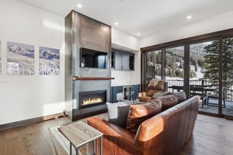 A cozy living room in Last Chair Lair with a brown leather sofa, a modern fireplace, a wet bar, and large glass doors leading to a balcony with mountain views.