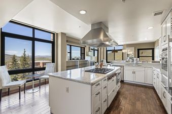 A contemporary kitchen featuring white cabinetry, light granite countertops, a large island with a gas cooktop, and stainless steel appliances, all set against a backdrop of expansive windows with mountain views.