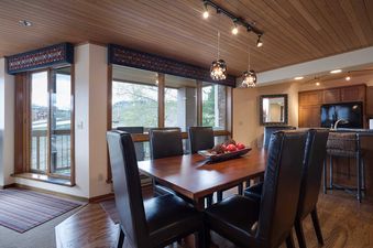 The dining area at Chateau Chamonix CX132 features a wooden table, black leather chairs, and two unique pendant lights, with large windows overlooking the outside scenery.