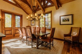 A rustic dining room with a large wooden table, upholstered chairs, a chandelier, and windows looking out onto autumn trees.