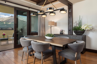 A dining room with a view. The dining room is furnished with a large wooden table, six chairs, and a large pendant light. The dining room is located in Lumiere One Bedroom Deluxe Residence, where guests can enjoy stunning views of the mountain peaks.