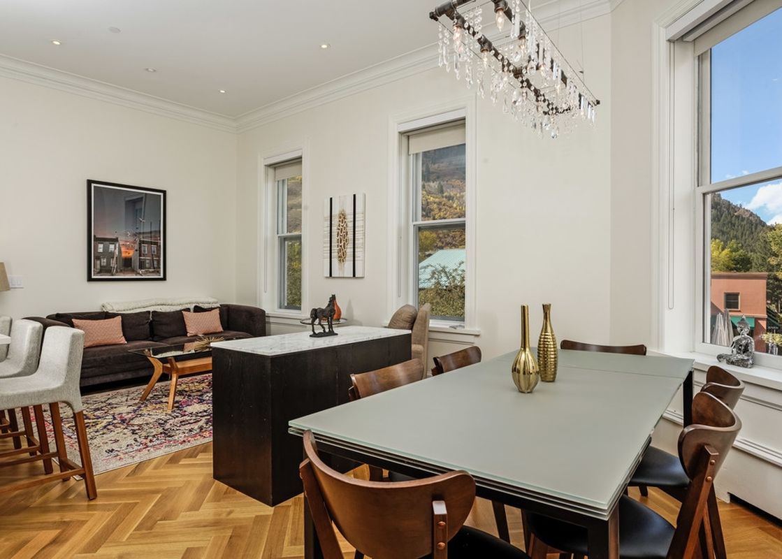 A large living room with a dining room and kitchen. There is a large couch with dark brown upholstery and a modern coffee table in the middle of the room. The dining room table is made of glass and has a modern feel. The room has large windows with a view