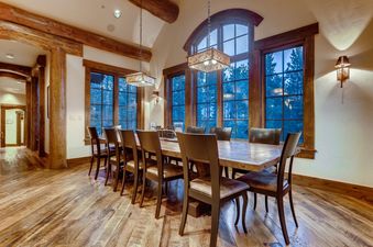 The dining area in Ski Hi Lodge features a long wooden table with multiple chairs, large windows revealing a forest, and decorative overhead lighting fixtures.