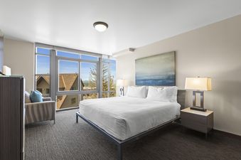 A modern bedroom with a king-sized bed, white linens, two bedside tables with lamps, and a large abstract painting above the bed, with expansive windows offering a view of houses and mountains.