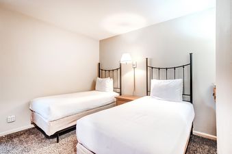 The secondary bedroom at Snow Flower 150 Park City showcases two twin beds with white bedding, black metal headboards, a wooden nightstand, and a wall lamp, all set against light-colored walls and speckled carpeting.