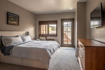 A cozy bedroom in Alpenglow Condo 26D features a king-sized bed, a wooden dresser, a window, and a glass door that opens to a mountain view.