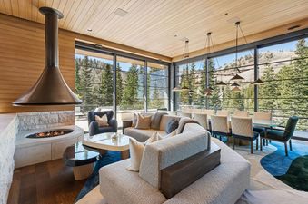 A spacious great room at Monitors Rest featuring a modern fireplace, a comfortable sectional sofa, a dining area, and large windows offering panoramic views of the surrounding mountains and pine trees.
