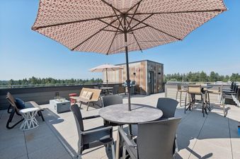 A spacious rooftop terrace at Columbia Crest 301 with various seating arrangements, a fire pit, and a BBQ grill under large patio umbrellas, offering clear views of the distant tree line and sky.