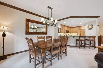 A rustic dining table with six chairs is positioned in the center of a well-lit room, with a view of a kitchen and breakfast bar in the background.