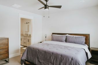 A modern bedroom with a comfortable bed featuring gray bedding and pillows, a dark wood headboard, and a stylish ceiling fan, with an open doorway leading to a well-lit bathroom.