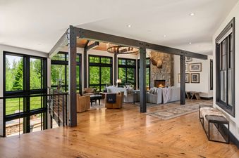 An expansive great room in a luxury mountain lodge with hardwood floors, timber beams, a stone fireplace, and large windows looking out onto a green forest.