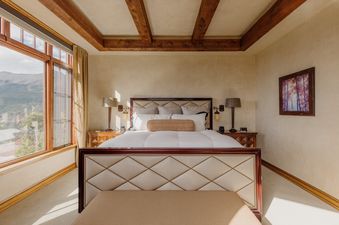 A luxurious bedroom features an upholstered bed, elegant nightstands, exposed wooden ceiling beams, and a large window offering panoramic mountain views, with an abstract tree painting on the wall.