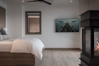 A modern bedroom in Riverlight Lodge features a large bed with neutral bedding, a wall-mounted TV displaying an airplane scene, and a stylish fireplace with glowing embers.