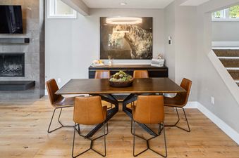 A modern dining room featuring a dark wood table with brown leather chairs, a circular chandelier, and a large horse painting above a credenza, with a fireplace and stairs visible.