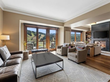 A spacious living room with a large leather sofa, two chairs, a coffee table and a fireplace with a mountain view out of the large windows.