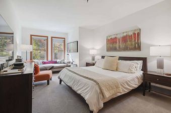 A spacious bedroom features a large bed with a tan quilted blanket, white linens, and a tan throw pillow, a dresser to the left, and a nightstand with a lamp to the right.  A small couch is located to the left of the bed, and two large windows with views