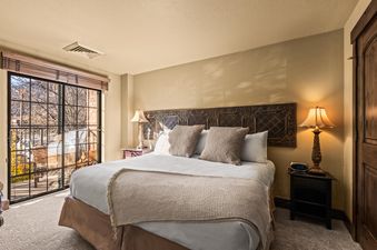 A cozy bedroom at Copperbottom Inn 103 features a large bed with a white duvet, decorative pillows, a textured throw, and an ornate metal headboard, beside a nightstand with a lamp and digital clock.