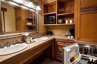 Double-sink vanity with tiled countertop, wooden cabinets, microwave, and open mini-fridge stocked with drinks in a hotel suite.