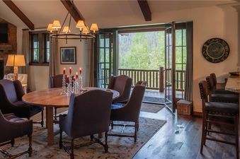 The dining area at Martin Creek Cottage features a round wooden table, dark upholstered chairs, a chandelier, and access to a balcony with forest views.