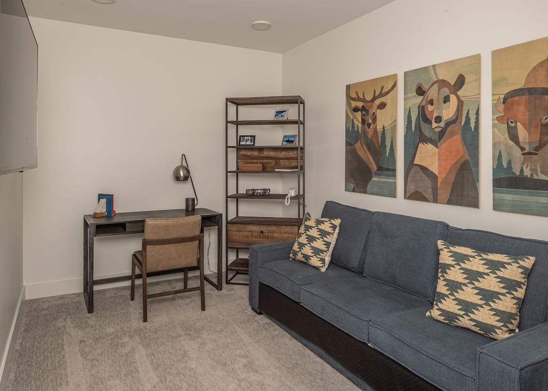 A modern and cozy den area features a blue sofa, a dark wooden desk with a leather chair, a tall metal bookshelf, and three abstract animal portraits on the wall.