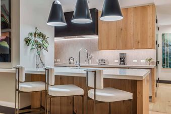 A modern kitchen with light wood cabinetry, white countertops, black pendant lights, and three white bar stools at the island.