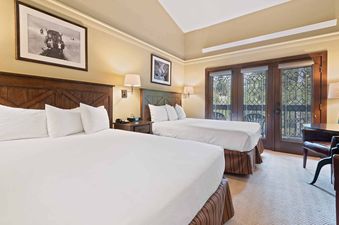 A spacious bedroom at The Charter C2105 features two beds with white linens, dark wood headboards, and a nightstand with a lamp, telephone, and alarm clock between them. Two framed pictures adorn the wall above each bed, and decorative glass double doors