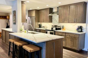 A modern kitchen with dark wood cabinets, white quartz countertops, stainless steel appliances, and a kitchen island with three bar stools.