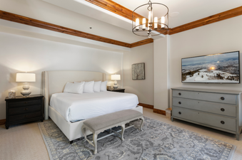 Grand Lodge Five Bedroom 402 features a king-size bed with a gray and white patterned rug, white linens, and a plush bench at the foot of the bed.