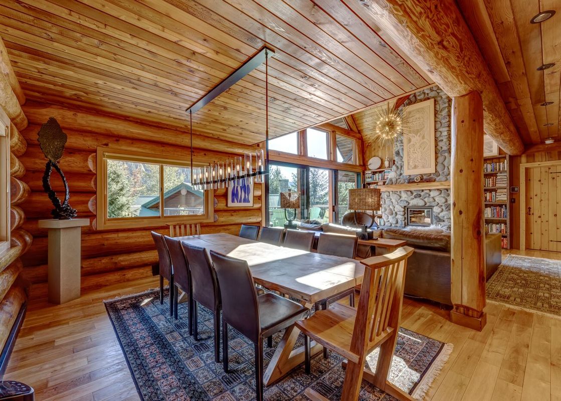 A spacious, light-filled dining area in a log cabin, featuring a large wooden table with ten chairs, a modern chandelier, and a view of the outdoors through the windows.