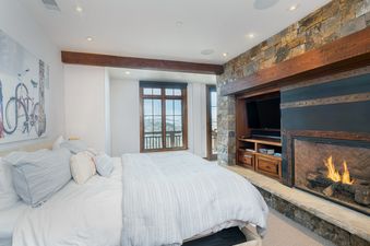 A cozy bedroom at See Forever San Sophia 114 3BR 4BA featuring a large bed, a fireplace built into a stone wall with a television above, and a window offering views of snowy mountains.
