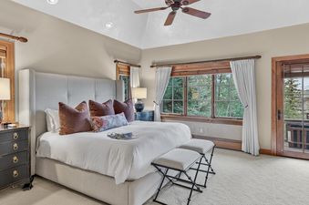 The primary bedroom at Timber Haven features a large bed with a tall upholstered headboard, nightstands with lamps, and large windows offering views of lush greenery and distant mountains.