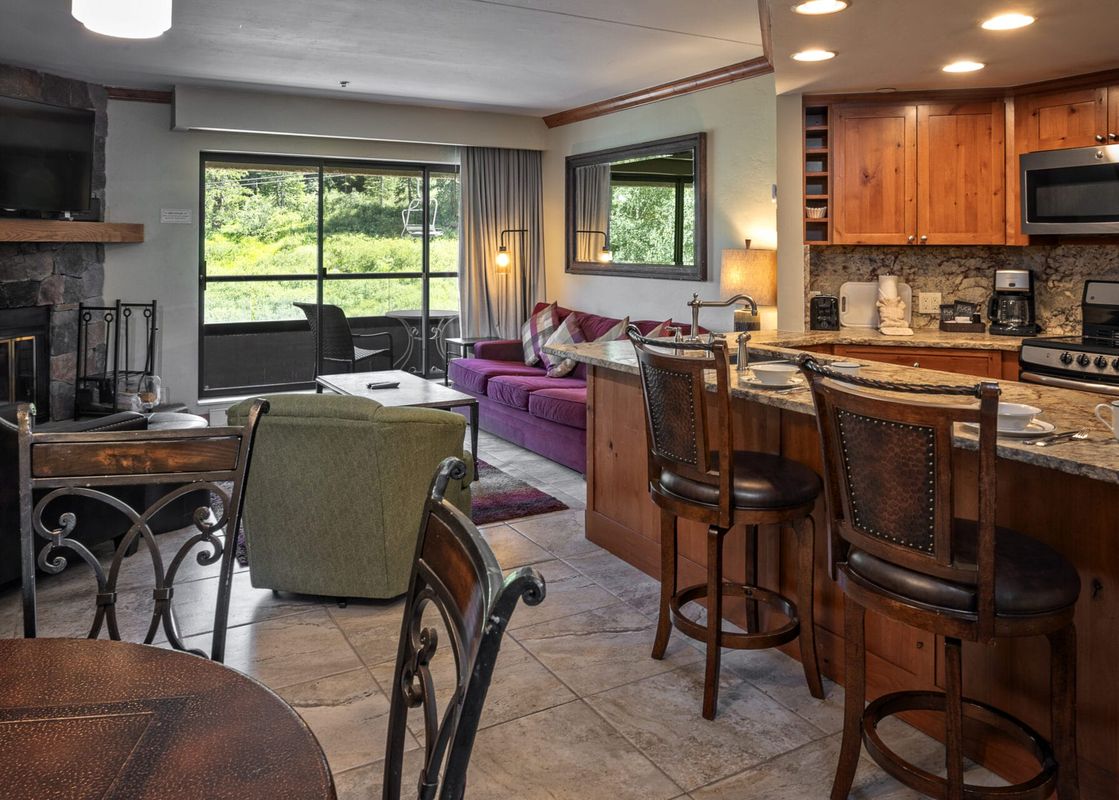 An inviting open-concept living space in Building 4 One Bedroom Beaver Run Resort features a purple sofa, a dining area, and a fully equipped kitchen with a breakfast bar, all overlooking a lush green exterior through a large glass door.