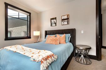 A bedroom with a plush bed with a light blue duvet, a throw rug at the end of the bed and a decorative pillow on the bed. there is a nightstand to the right with an oval metal table.